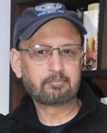 ashok pande | No. 1 Indian Media News Portal Portrait of a man wearing a dark cap and glasses, with a short beard, standing in front of a bookshelf.