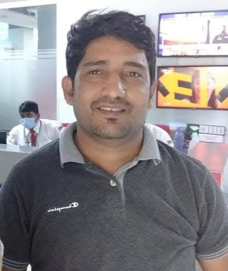 awdhesh yadav | No. 1 Indian Media News Portal Man with dark hair and stubble wearing a gray polo shirt, standing in an office with a masked coworker in the background.
