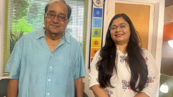 Two smiling people stand side by side in a sunny room: an older man in a blue patterned shirt on the left and a young woman with long dark hair and glasses on the right, with a window and colorful decor behind them.