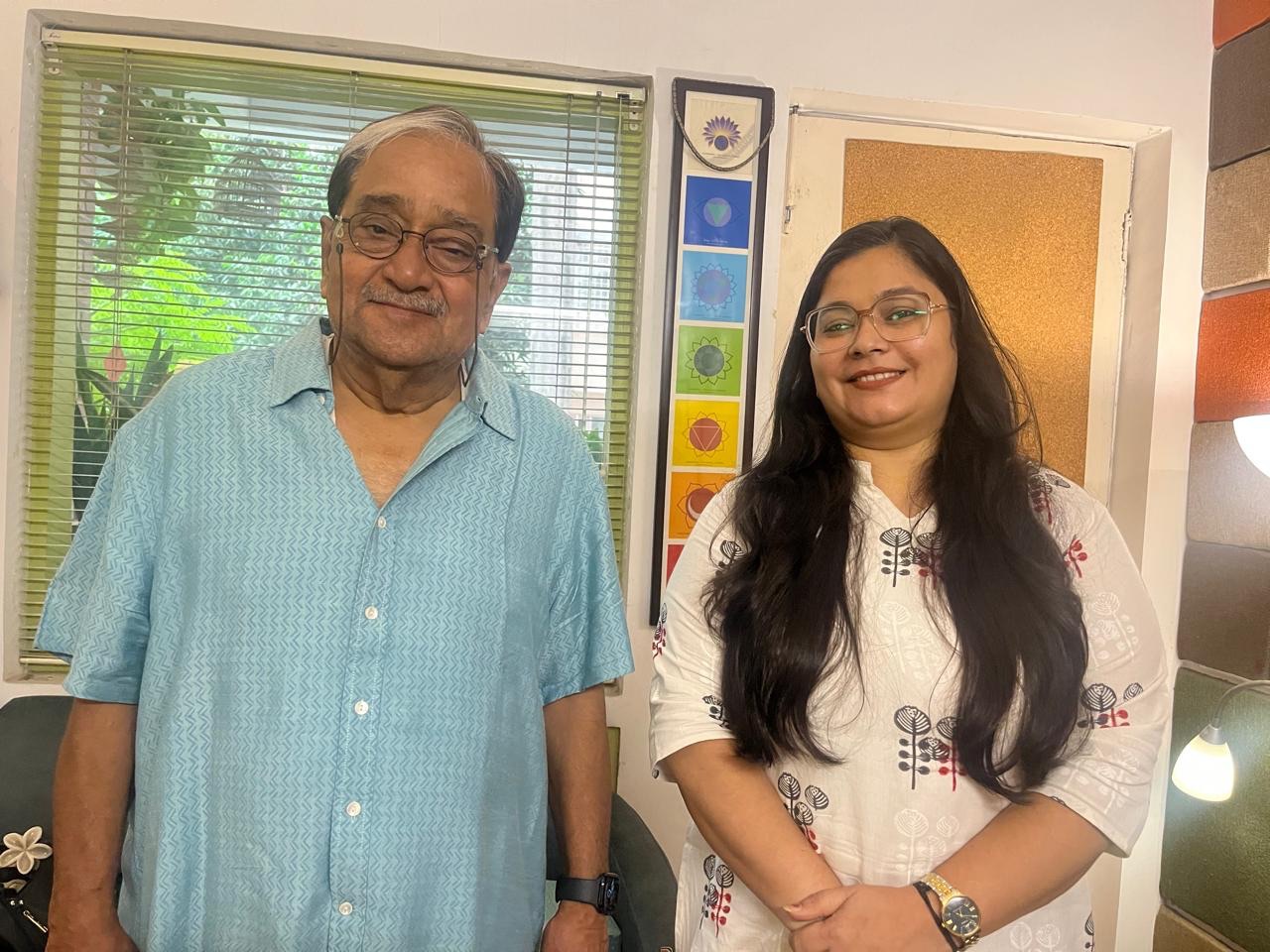 Two smiling people stand side by side in a sunny room: an older man in a blue patterned shirt on the left and a young woman with long dark hair and glasses on the right, with a window and colorful decor behind them.
