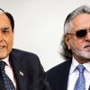 Two mature men in business suits posing for a photo; left man wears a pink shirt and Indian flag pin, right man wears sunglasses and a navy suit.