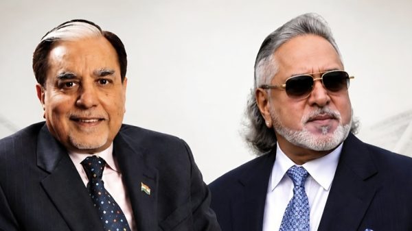 Two mature men in business suits posing for a photo; left man wears a pink shirt and Indian flag pin, right man wears sunglasses and a navy suit.