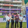 Four men pose together on a grassy cricket stadium field, with colorful banners and seating stands in the background.