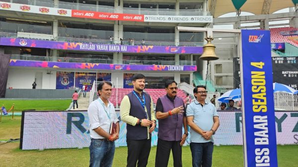 Four men pose together on a grassy cricket stadium field, with colorful banners and seating stands in the background.