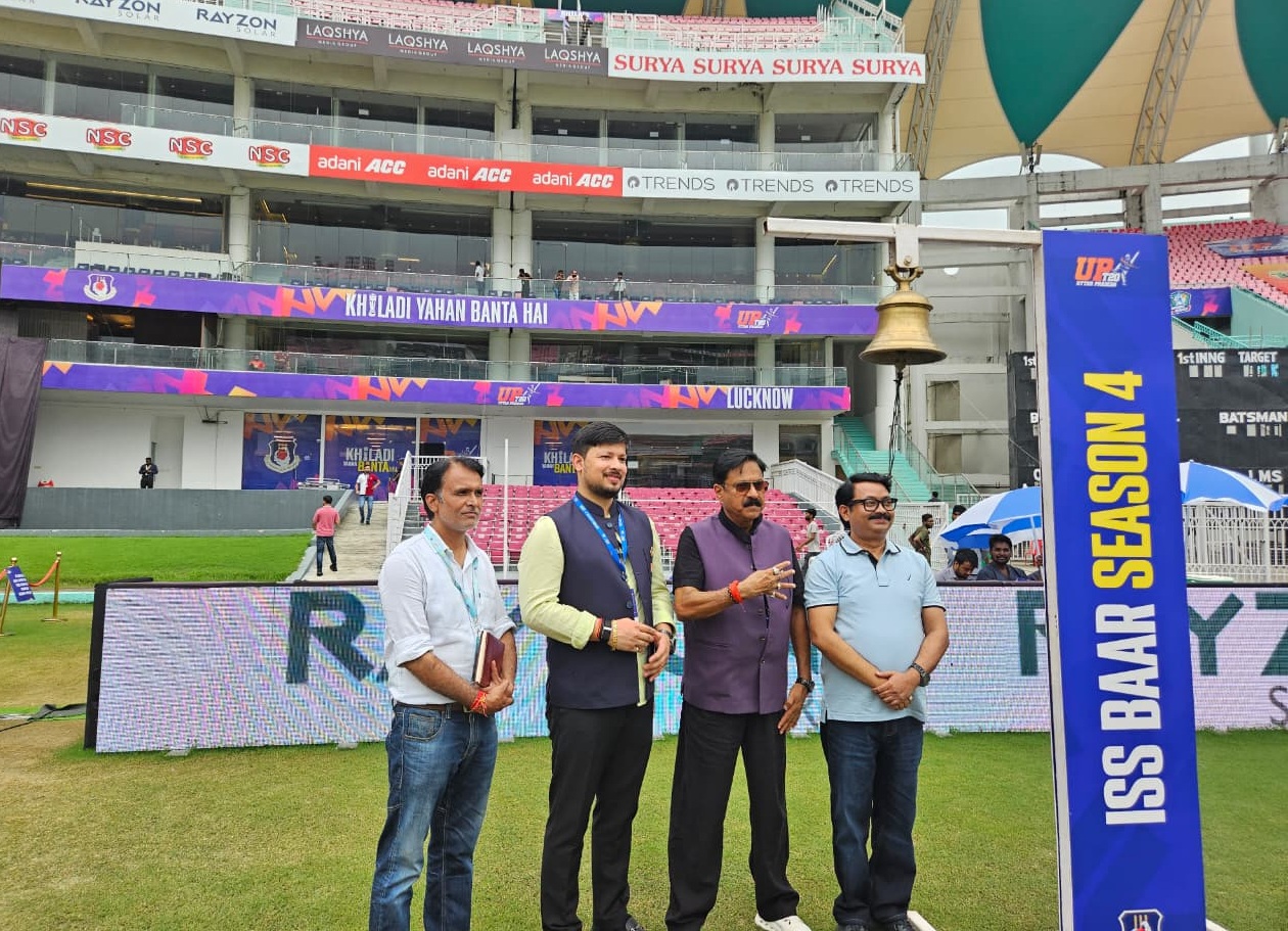 Four men pose together on a grassy cricket stadium field, with colorful banners and seating stands in the background.