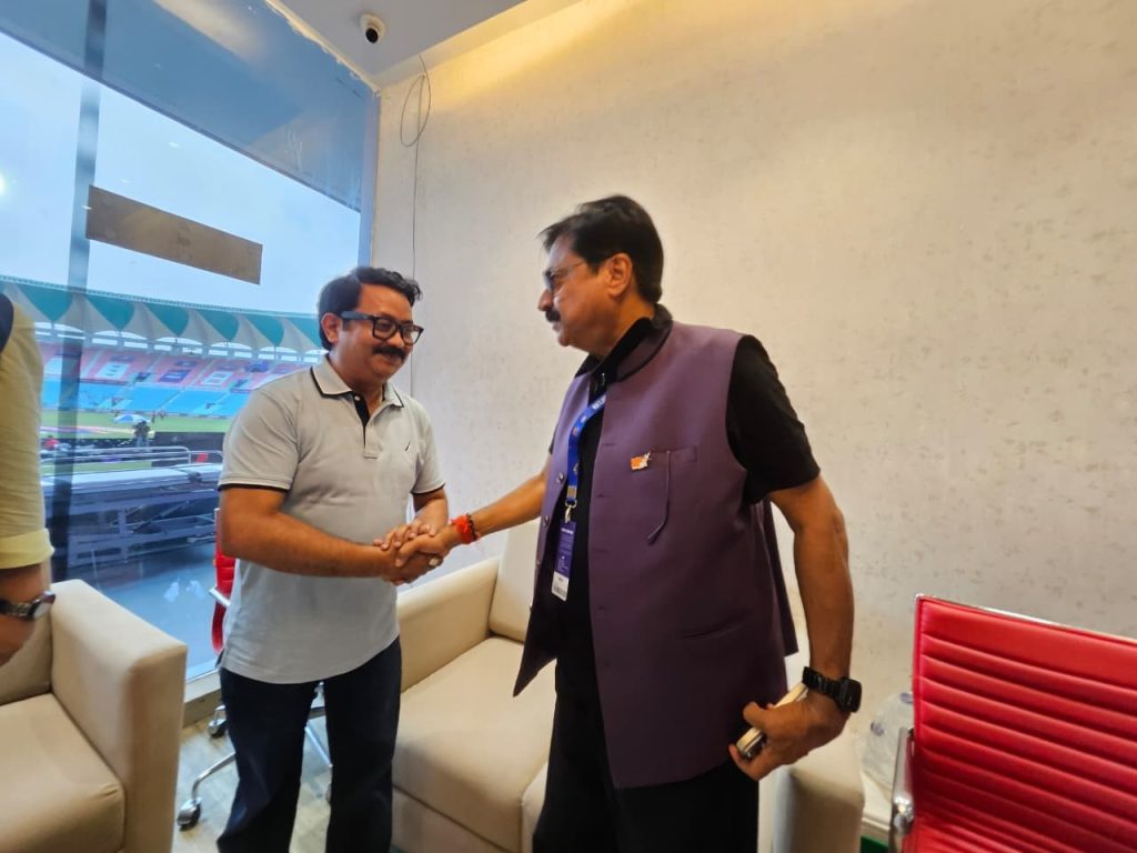 chandrasen_2 | No. 1 Indian Media News Portal Two men shake hands in a modern room, a stadium visible through a large window behind them.