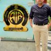 Man in a dark shirt and beige pants standing beside a blue-green wall with a circular emblem and Devanagari text outdoors in front of a signboard.