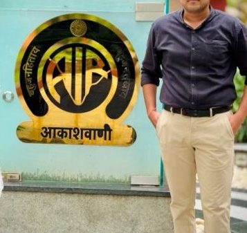 Man in a dark shirt and beige pants standing beside a blue-green wall with a circular emblem and Devanagari text outdoors in front of a signboard.