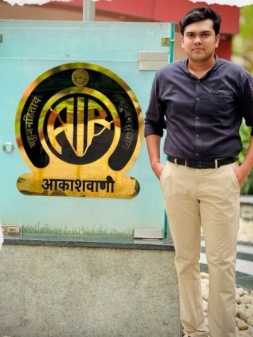 Man in a dark shirt and beige pants standing beside a blue-green wall with a circular emblem and Devanagari text outdoors in front of a signboard.
