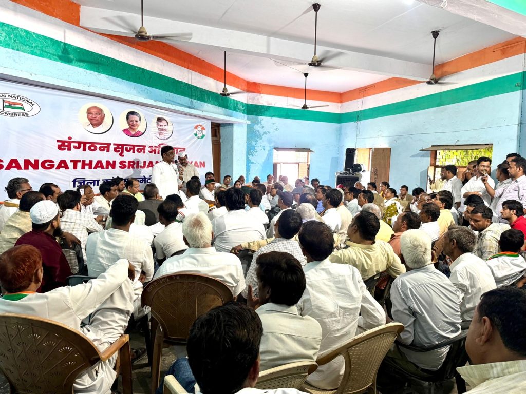 congress_bs | No. 1 Indian Media News Portal Community meeting with a speaker addressing a crowded hall, banner behind reading a political party name and portraits on the wall.