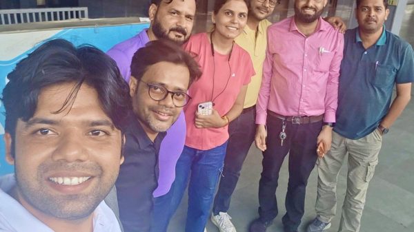 Group of seven people posing for a selfie outdoors, all smiling at the camera in a sunny courtyard area outside a building.