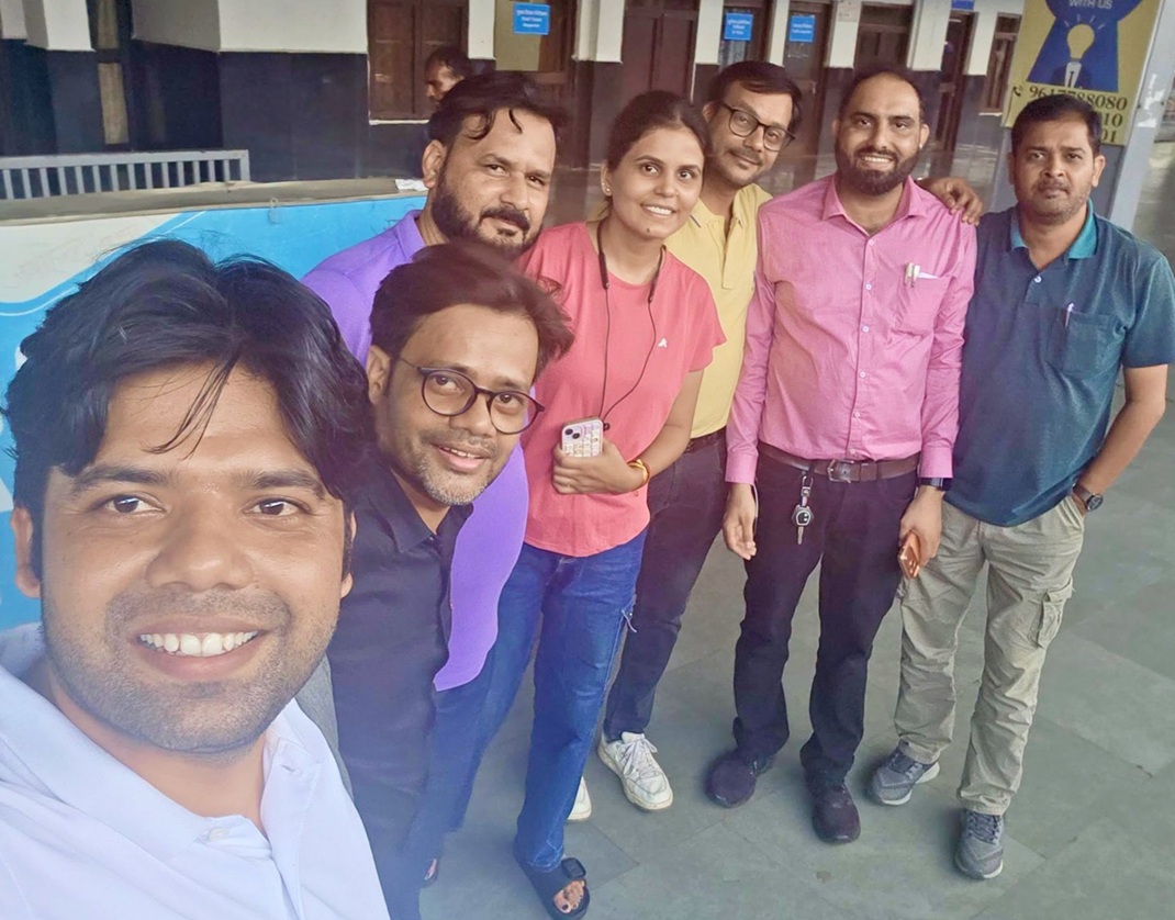 Group of seven people posing for a selfie outdoors, all smiling at the camera in a sunny courtyard area outside a building.