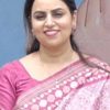 Portrait of a smiling woman in a pink saree standing against a blue wall behind her.
