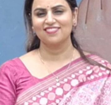 Portrait of a smiling woman in a pink saree standing against a blue wall behind her.