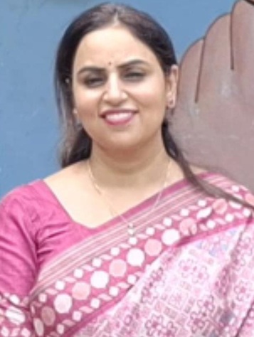 Portrait of a smiling woman in a pink saree standing against a blue wall behind her.