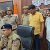 Indian police officers in khaki uniforms stand behind a seated officer at a press briefing; Indian flag and badges visible on the wall behind them.
