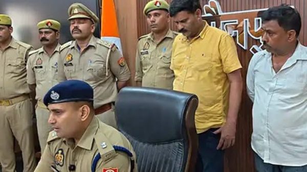 Indian police officers in khaki uniforms stand behind a seated officer at a press briefing; Indian flag and badges visible on the wall behind them.