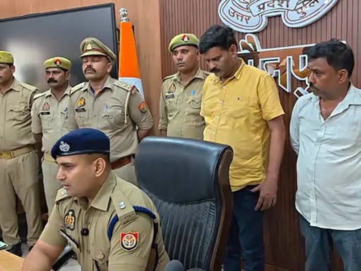 Indian police officers in khaki uniforms stand behind a seated officer at a press briefing; Indian flag and badges visible on the wall behind them.