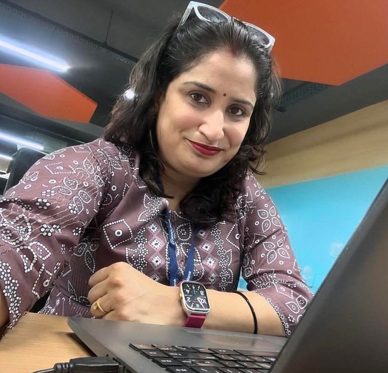 divya mishra | No. 1 Indian Media News Portal Woman sitting at a desk with a laptop, wearing a maroon patterned top, glasses on her head, a pink smartwatch, and a lanyard, smiling at the camera in an office setting.