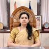 Woman in a beige sari sits at a desk in a formal office with awards and a framed photo behind her.
