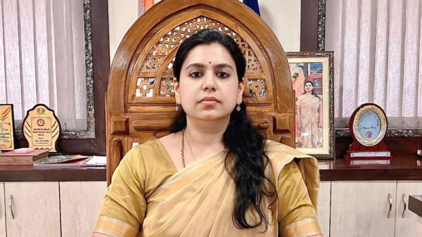 Woman in a beige sari sits at a desk in a formal office with awards and a framed photo behind her.