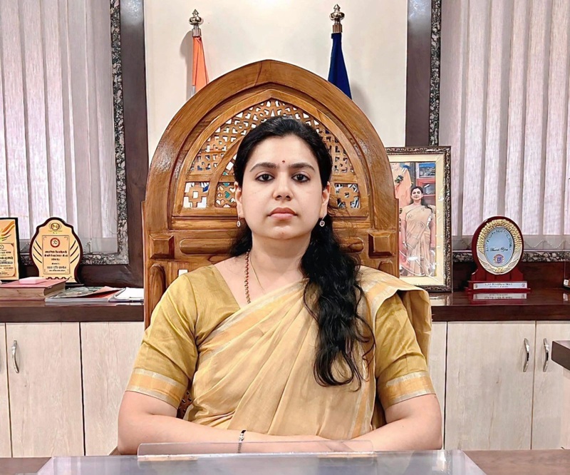 Woman in a beige sari sits at a desk in a formal office with awards and a framed photo behind her.