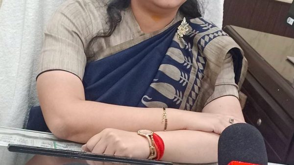 Woman in a blue sari sitting at a desk with microphones, likely giving a press statement.