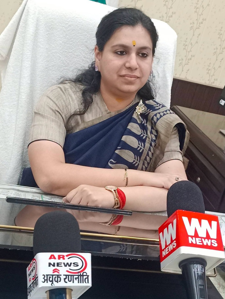 Woman in a blue sari sitting at a desk with microphones, likely giving a press statement.