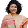 Smiling woman with curly black hair, wearing a pink and beige sari with a bindi, against a white background.