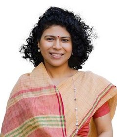Smiling woman with curly black hair, wearing a pink and beige sari with a bindi, against a white background.