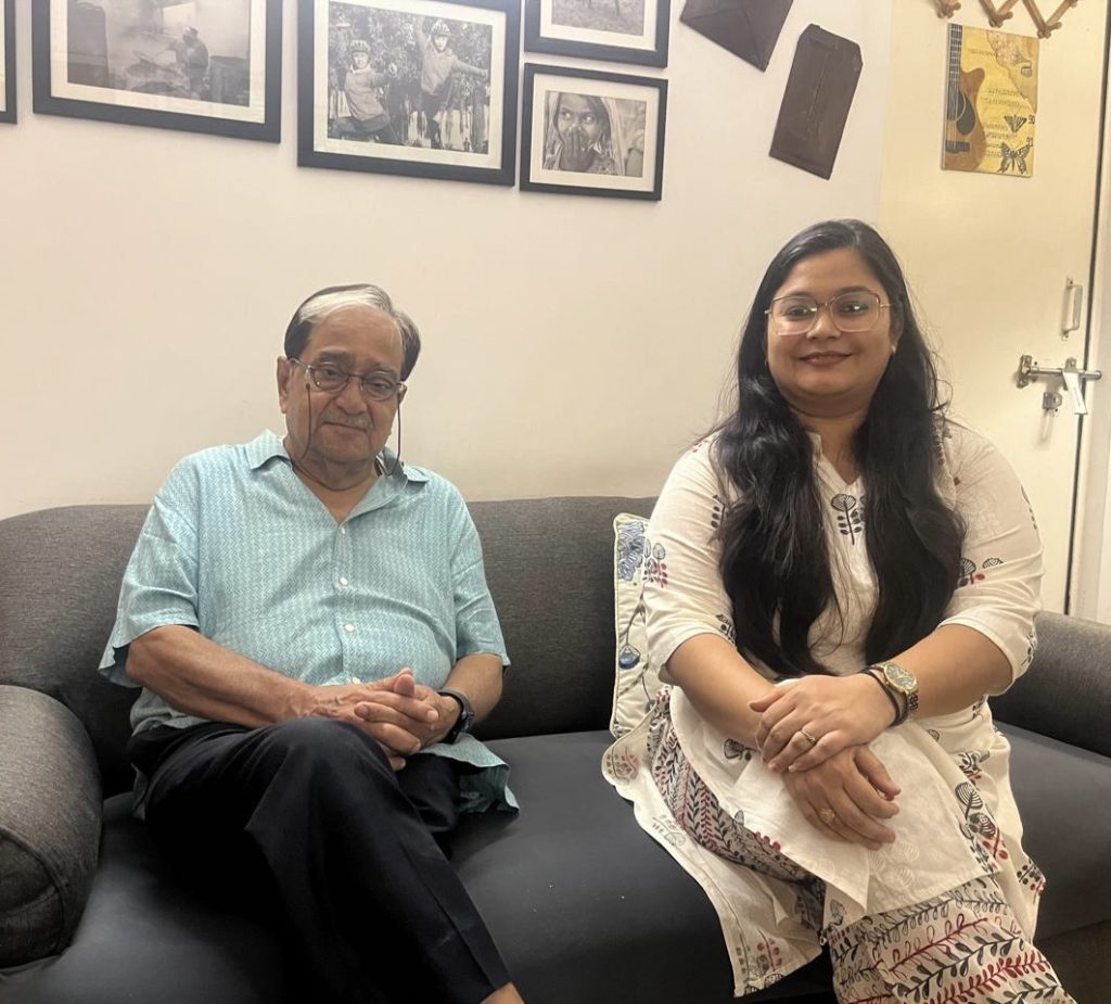 e52b933c-7cd6-4d86-b5aa-2819ae049877 | No. 1 Indian Media News Portal Older man and younger woman sit on a gray sofa in a living room with framed photos on the wall behind them.