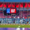 Cricket stadium at dusk with a large LED screen showing Hindi text 'आज की खबर' and bright stage lights in the stands around it, mostly empty seating.