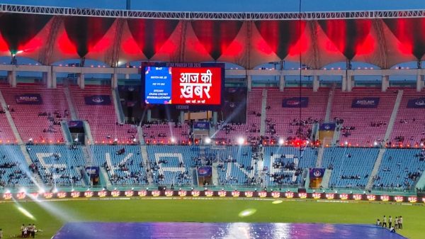 Cricket stadium at dusk with a large LED screen showing Hindi text 'आज की खबर' and bright stage lights in the stands around it, mostly empty seating.