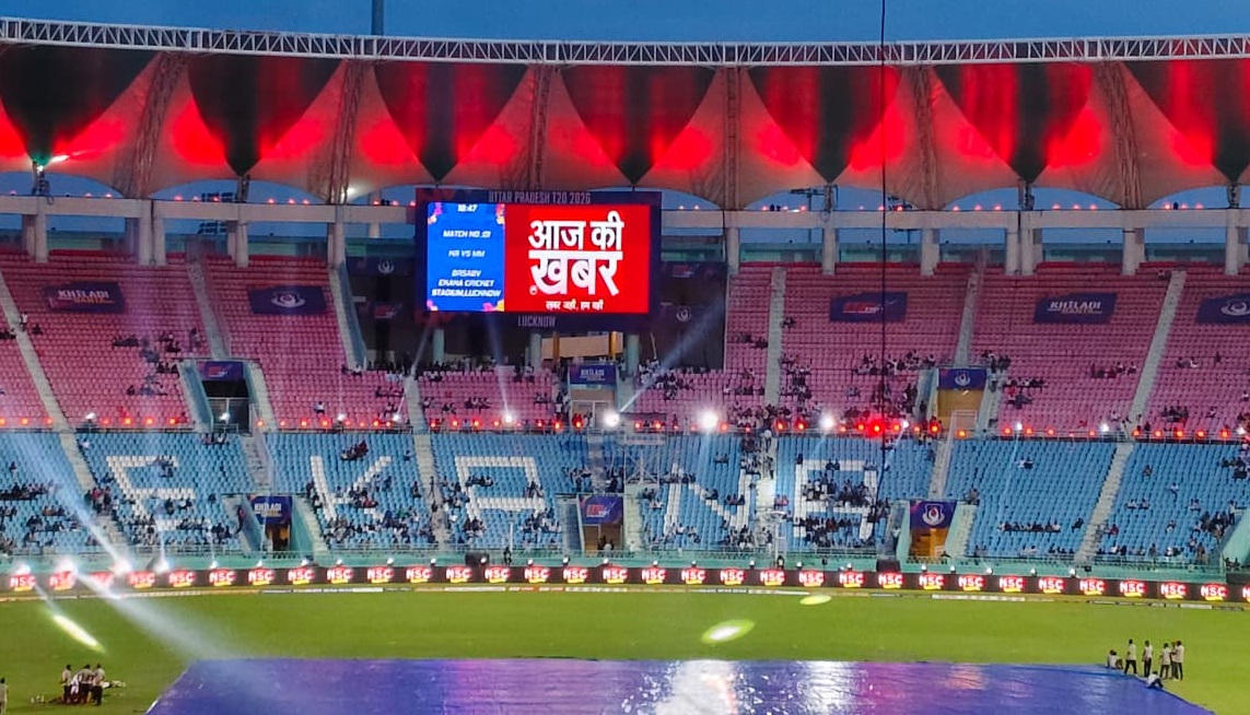 Cricket stadium at dusk with a large LED screen showing Hindi text 'आज की खबर' and bright stage lights in the stands around it, mostly empty seating.