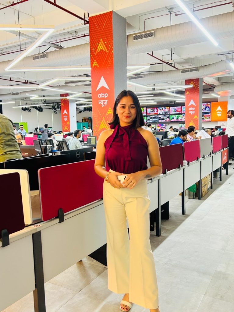 f5ffc395-5947-4d06-8d2b-6e797342b54f | No. 1 Indian Media News Portal Woman in a burgundy sleeveless top and beige wide-leg pants stands at a news desk in a busy ABP News office.