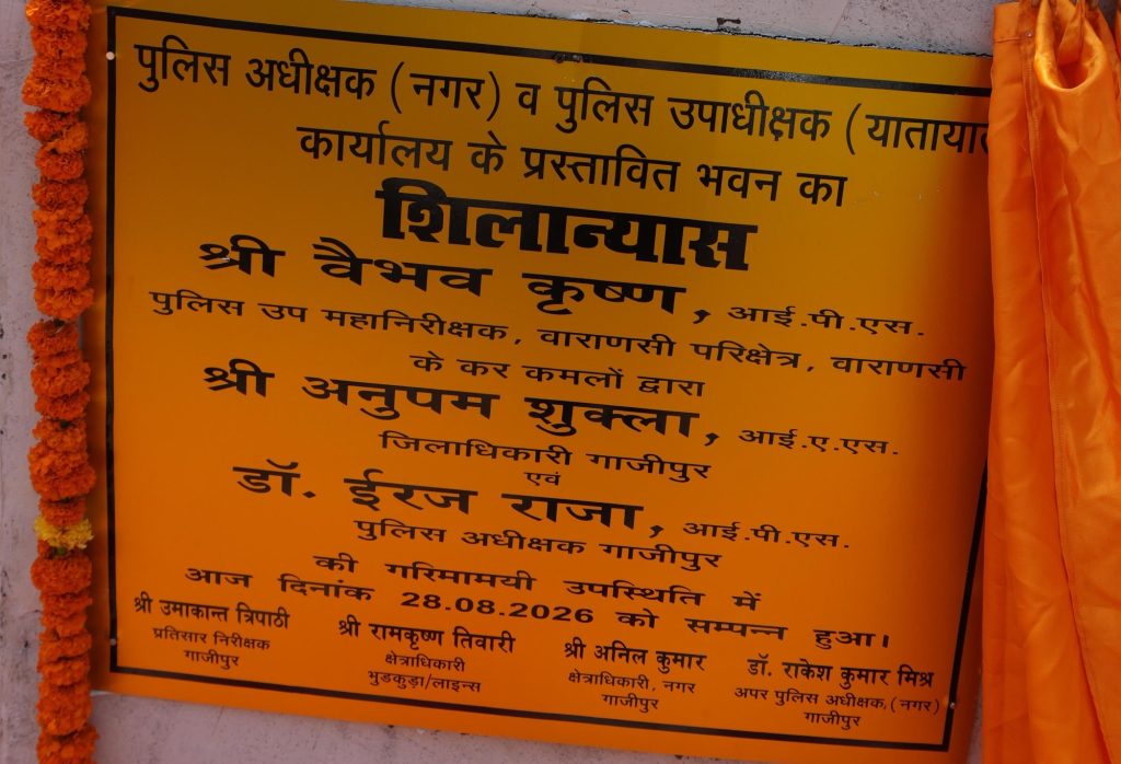 fd689d43-338e-4567-b7d6-efd37993ad22 | No. 1 Indian Media News Portal Yellow public notice plaque with black Hindi text, bordered in black, flanked by orange marigold garlands on the left and an orange curtain on the right.