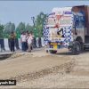 A large, decorated dump truck on a gravel road beside a striped concrete barrier, with several men watching a road construction site nearby.