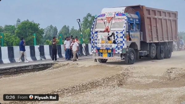A large, decorated dump truck on a gravel road beside a striped concrete barrier, with several men watching a road construction site nearby.