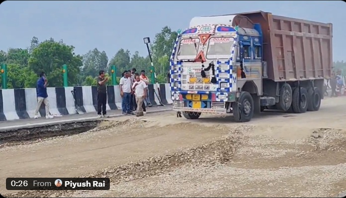 A large, decorated dump truck on a gravel road beside a striped concrete barrier, with several men watching a road construction site nearby.