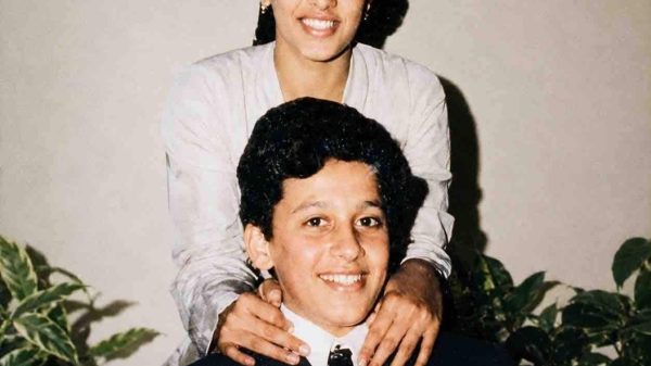 Young man in a dark suit sits smiling as a woman stands behind him with hands on his shoulders.