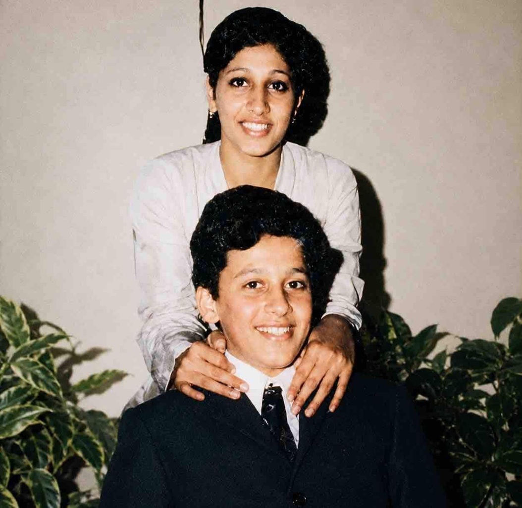 Young man in a dark suit sits smiling as a woman stands behind him with hands on his shoulders.