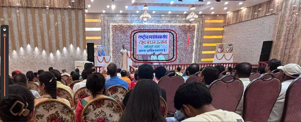 ghadhipuri | No. 1 Indian Media News Portal Indoor ceremony in a decorated hall with a stage and audience seated facing a speaker at a podium; a framed banner with Hindi text is centered on the stage background.