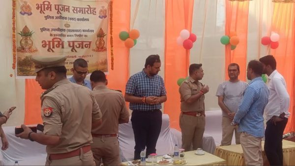 Group of police officers and civilians chatting under orange drapes at a Bhoomi Pujan ceremony with a Hindi banner in the background.