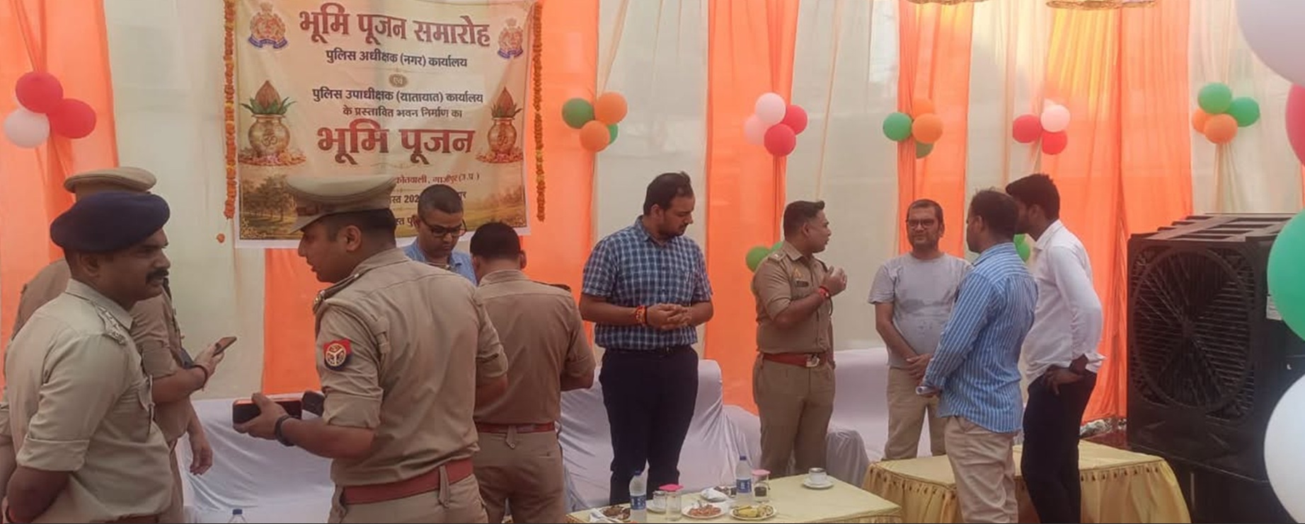 Group of police officers and civilians chatting under orange drapes at a Bhoomi Pujan ceremony with a Hindi banner in the background.