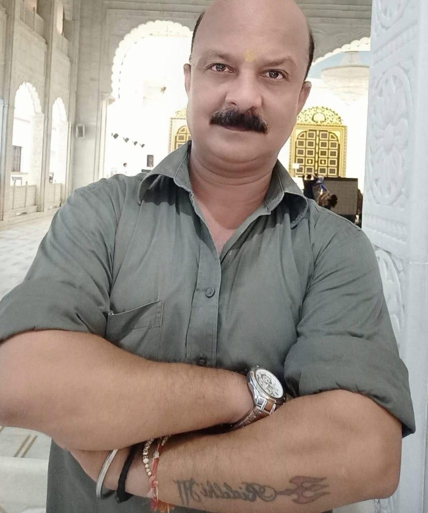 gyanu | No. 1 Indian Media News Portal Self-portrait of a man with a bald head and mustache, arms crossed, wearing a gray shirt inside a bright temple with ornate doors in the background.