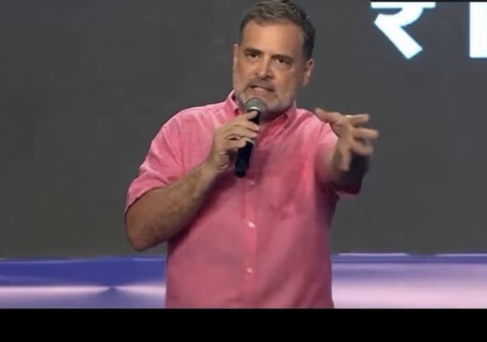 Man in a pink shirt speaks into a handheld microphone on stage, gesturing with his right hand.