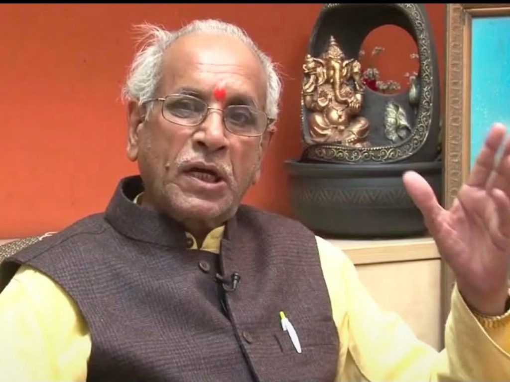 img_0391 | No. 1 Indian Media News Portal Older man with white hair and glasses wearing a yellow kurta and brown vest, speaking with his hand raised.