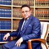 Man in a blue suit sitting in an office chair in front of shelves filled with law books, looking at the camera.