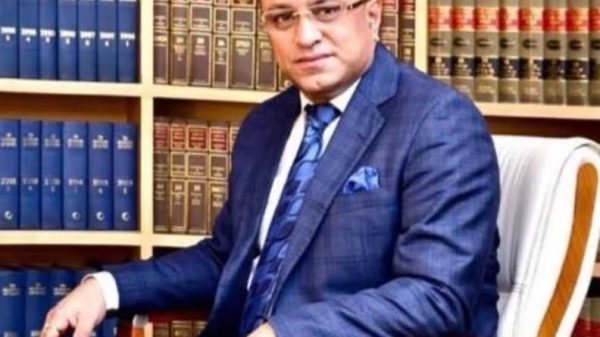 Man in a blue suit sitting in an office chair in front of shelves filled with law books, looking at the camera.