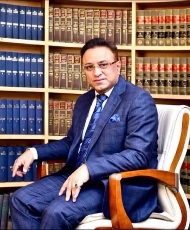 Man in a blue suit sitting in an office chair in front of shelves filled with law books, looking at the camera.